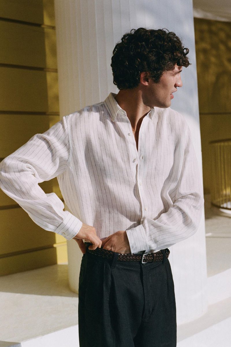 Model with dark curly hair wearing a white striped long-sleeve shirt and dark trousers against a pale columned building