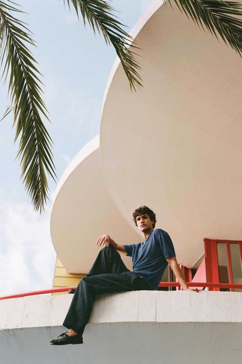 Model seated on a concrete ledge wearing a blue t-shirt and dark trousers beneath palm fronds.