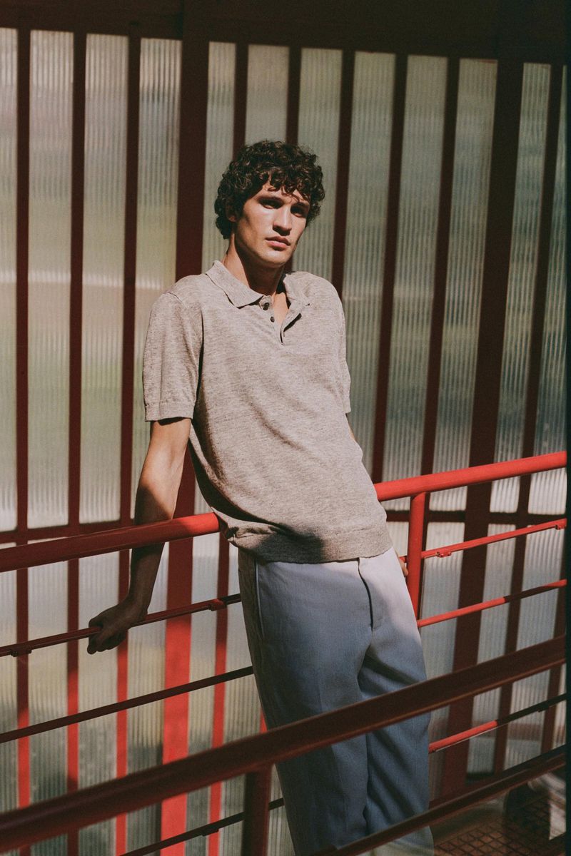 An adult male poses wearing a polo shirt and light blue trousers in front of a red and white partition with sunlight streaming through.