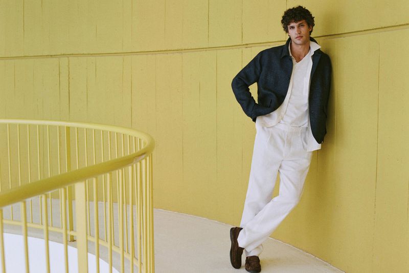 Model leaning against a yellow wall wearing a dark jacket, white shirt, and white trousers with brown loafers