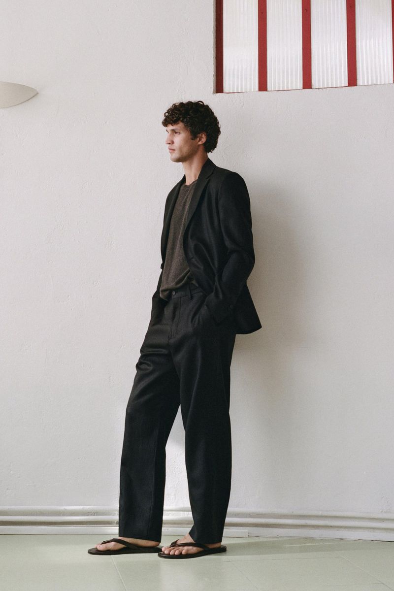 Full-length portrait of a male model wearing a dark blazer, trousers, and sandals against a white wall.