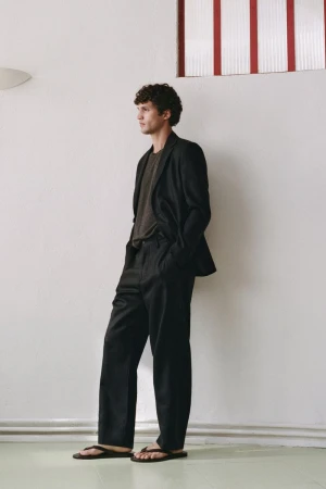 Full-length portrait of a male model wearing a dark blazer, trousers, and sandals against a white wall.