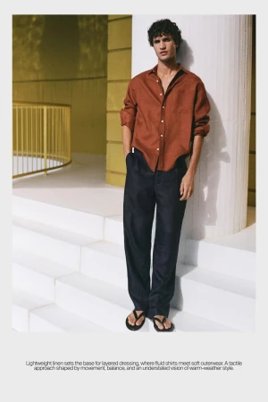 Man wearing a rust orange long-sleeve shirt and dark trousers leaning against a white pillar on stone steps