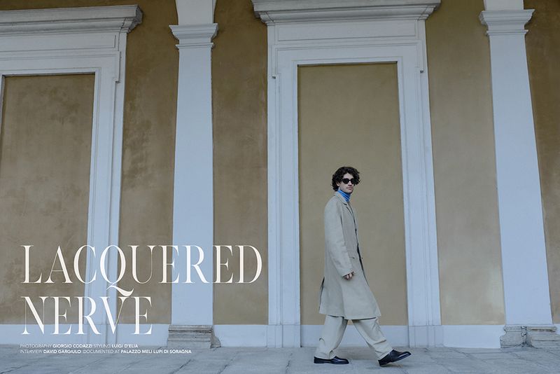 Man walking in profile wearing a beige trench coat and dark sunglasses before a mustard-yellow architectural wall with white columns