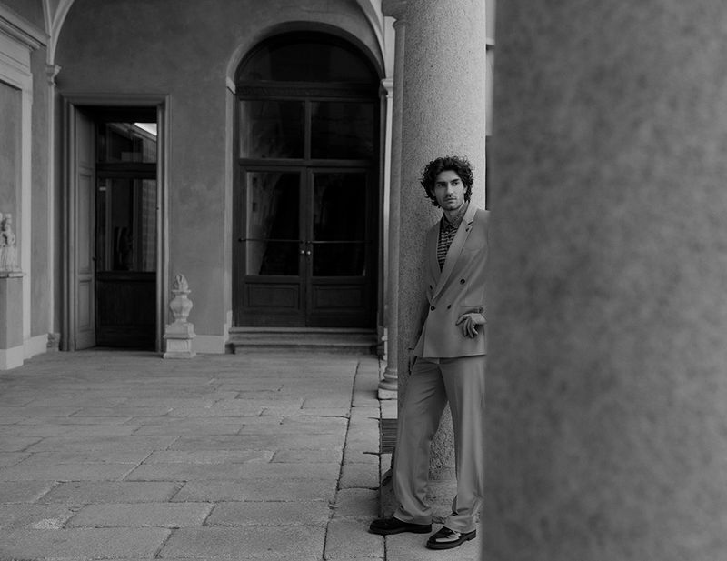Man wearing a double-breasted suit leaning against a classical stone column in an open courtyard for Vestal Magazine.