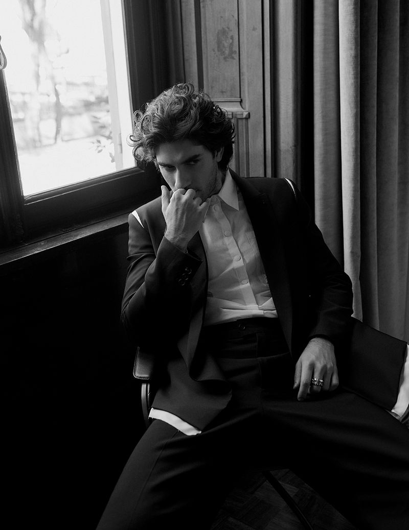 Man with curly dark hair wearing a dark suit jacket and white shirt seated indoors near a window for Vestal Magazine.