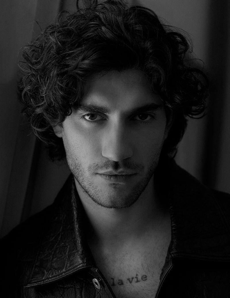 Black and white portrait of a man with dark, curly hair wearing a dark jacket and a tattoo on his chest, looking directly at the camera.