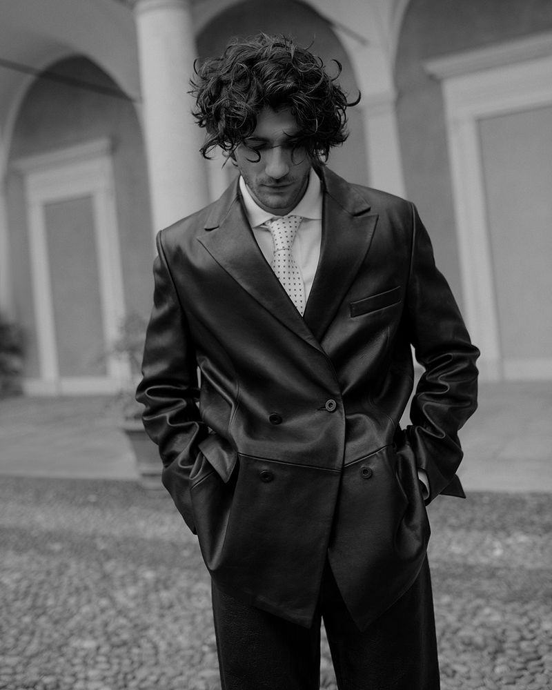 Man with curly dark hair wearing a dark double breasted suit jacket and tie standing outdoors