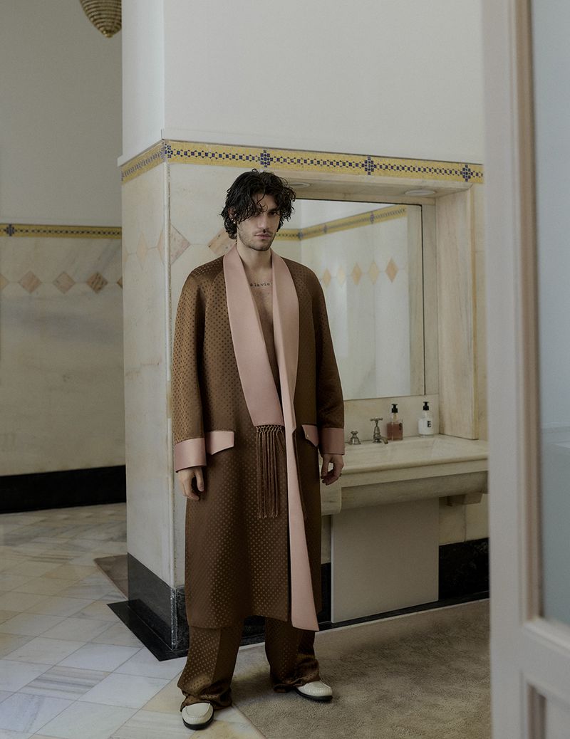 Man wearing a brown patterned robe with pink lapels standing in a marble-tiled bathroom for Vestal Magazine editorial