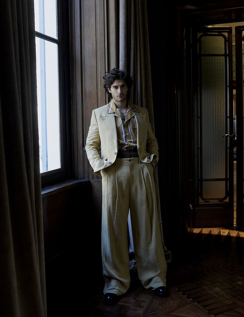 Man wearing a tailored yellow suit and striped shirt standing indoors next to a tall window with dark curtains.