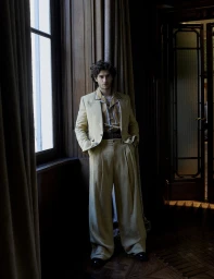 Man wearing a tailored yellow suit and striped shirt standing indoors next to a tall window with dark curtains.