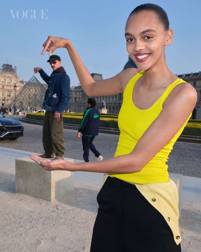 Model in a yellow tank top poses in the foreground, making it appear as though she is holding a smaller man standing in the distance.
