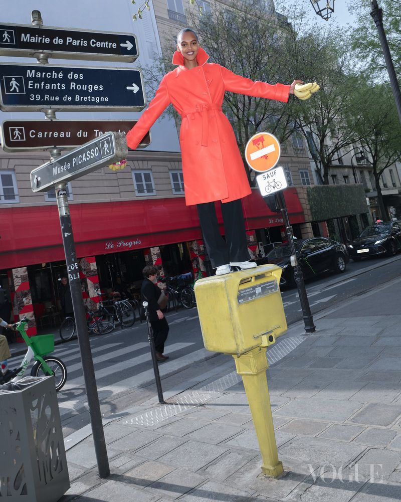 Model balancing on a yellow post in a bright orange coat holding bananas on a Parisian street with directional signs
