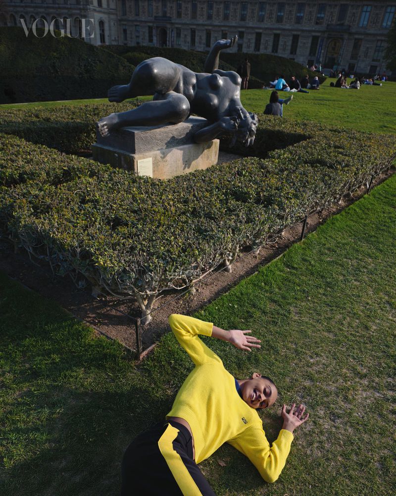 Model wearing yellow sweater lying on grass mimicking a bronze statue in a park.