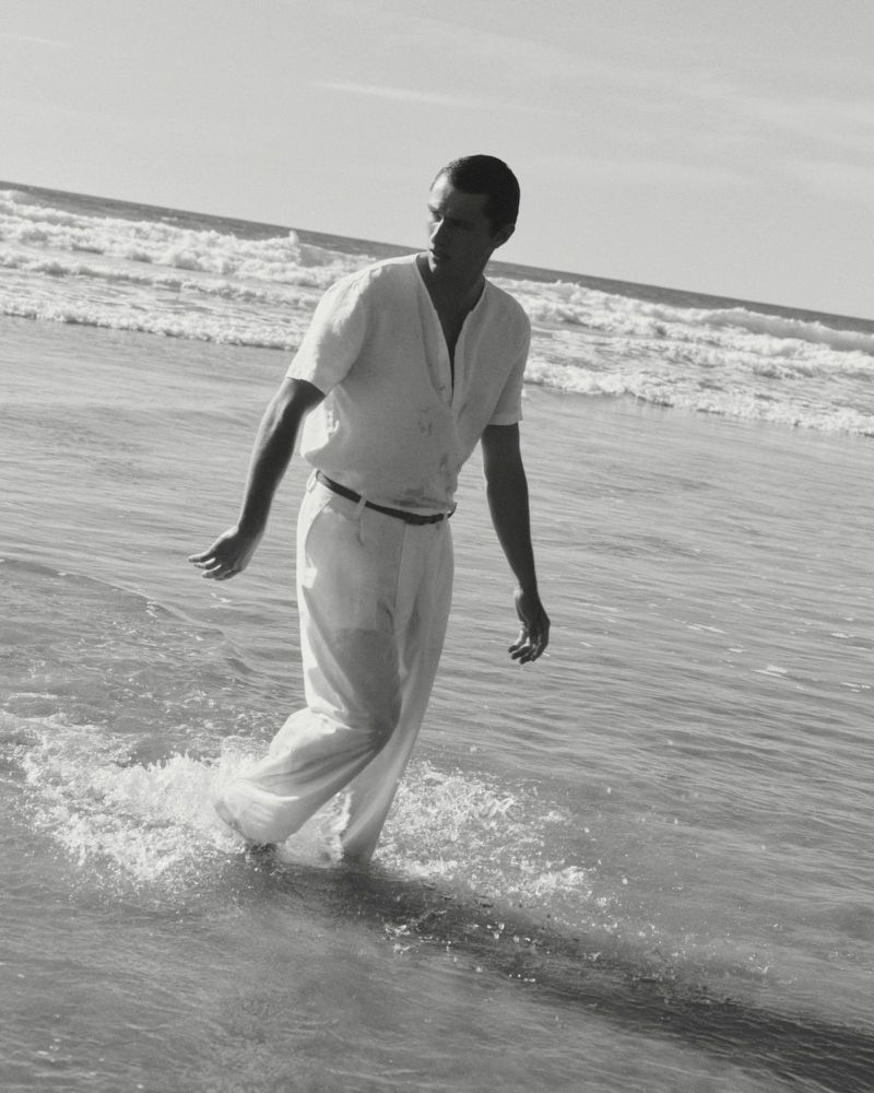 Man walking through shallow ocean water wearing a light shirt and trousers in a black and white beach scene.