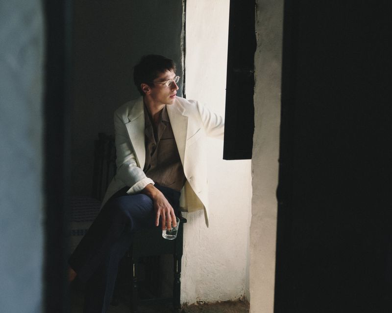 Man wearing a cream blazer and dark trousers holding a glass near an open window.