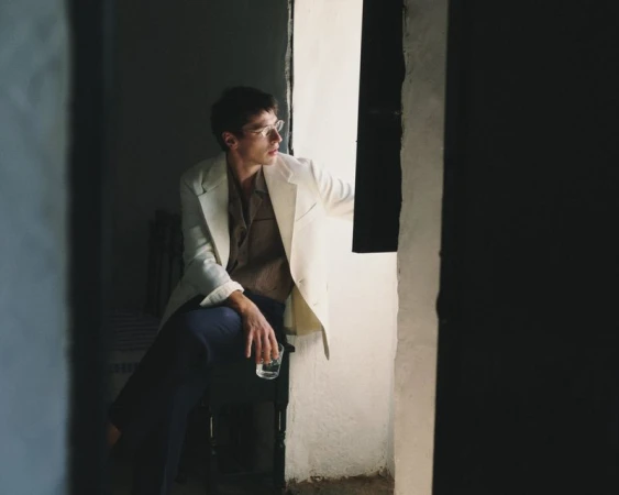 Man wearing a cream blazer and dark trousers holding a glass near an open window.