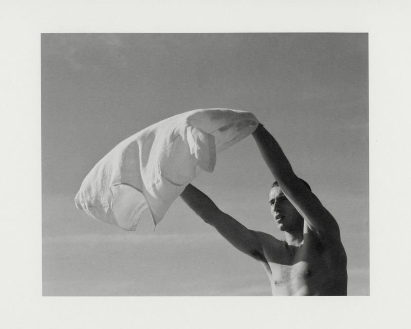 Monochrome portrait of a man holding a white shirt overhead against an open sky background
