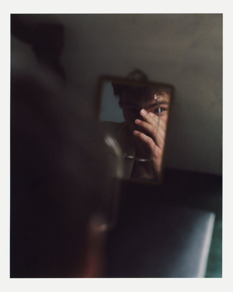 Close-up portrait of a young man looking into a small framed mirror reflecting his face.