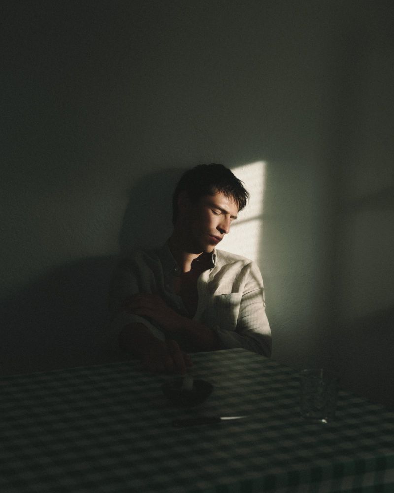 A man in a light-colored shirt seated at a checkered tablecloth table in a dimly lit room with a shaft of light.