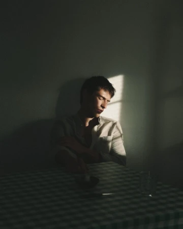 A man in a light-colored shirt seated at a checkered tablecloth table in a dimly lit room with a shaft of light.