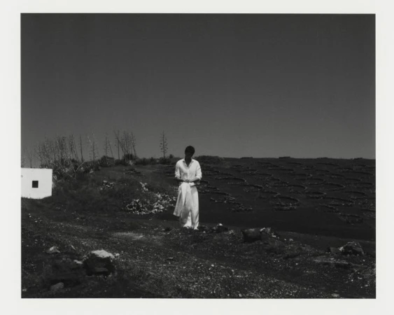 A man wearing a white shirt and trousers standing in a sparse outdoor landscape with dark volcanic terrain.