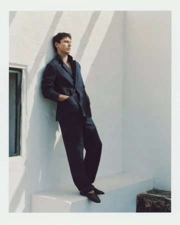 Male model wearing a dark navy blazer and trousers leaning against a sunlit white wall outdoors