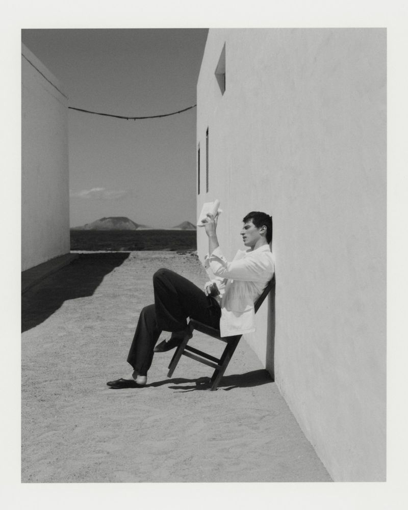 A man in a white jacket and dark trousers sitting on a tilted chair against a white wall outdoors.