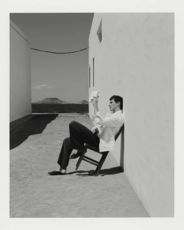 A man in a white jacket and dark trousers sitting on a tilted chair against a white wall outdoors.