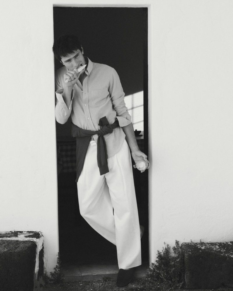 Man wearing button-down shirt and white trousers leaning in a doorway holding a small object