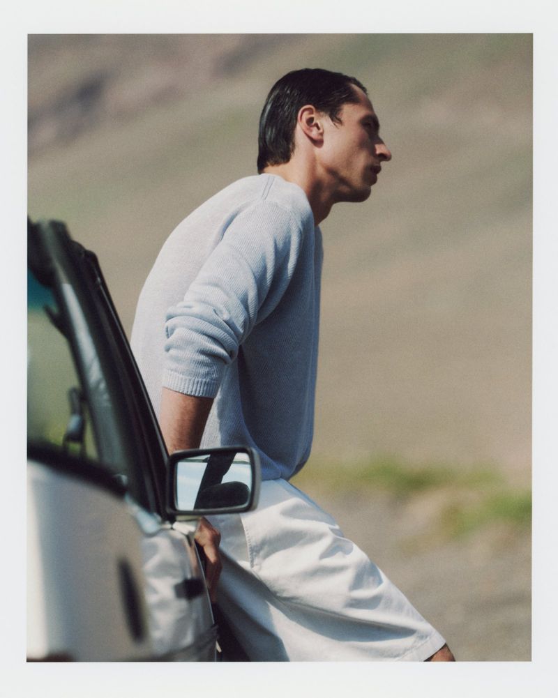 A man in a sweater and light pants leans against a car, with a blurred mountain background. The shot captures natural light and style.