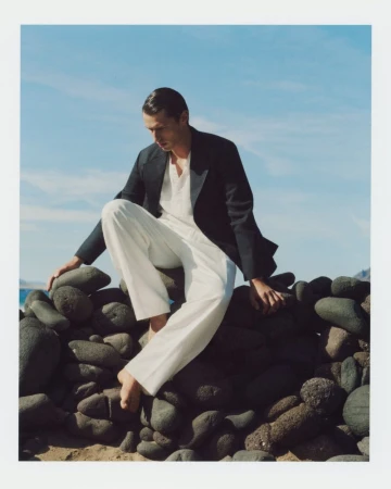 Man wearing a dark blazer and white trousers seated on dark rocks against a blue sky for Zara