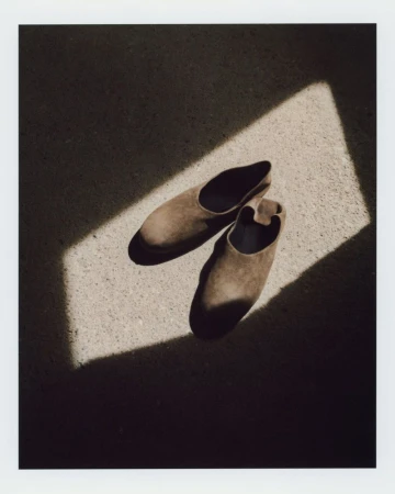 A pair of brown suede slip-on shoes rests on a textured concrete surface under a beam of bright sunlight.