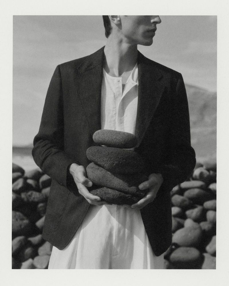 Man wearing a black blazer and white shirt holding a stack of smooth dark stones in an outdoor coastal setting.