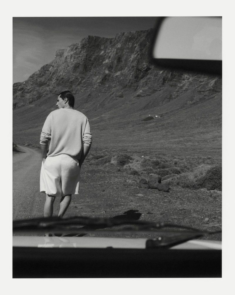 A man wearing a light sweater and shorts walking on a dirt road near mountains, shot from inside a car.