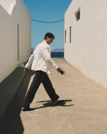 A man wearing a white jacket and dark trousers walking between two white buildings toward the ocean.