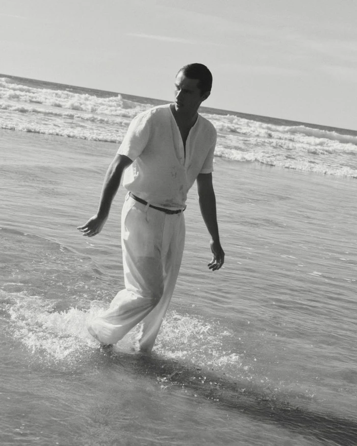Man walking through shallow ocean water wearing a light shirt and trousers in a black and white beach scene.