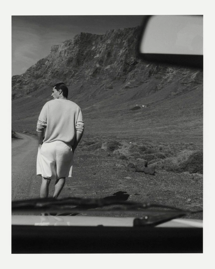 A man wearing a light sweater and shorts walking on a dirt road near mountains, shot from inside a car.