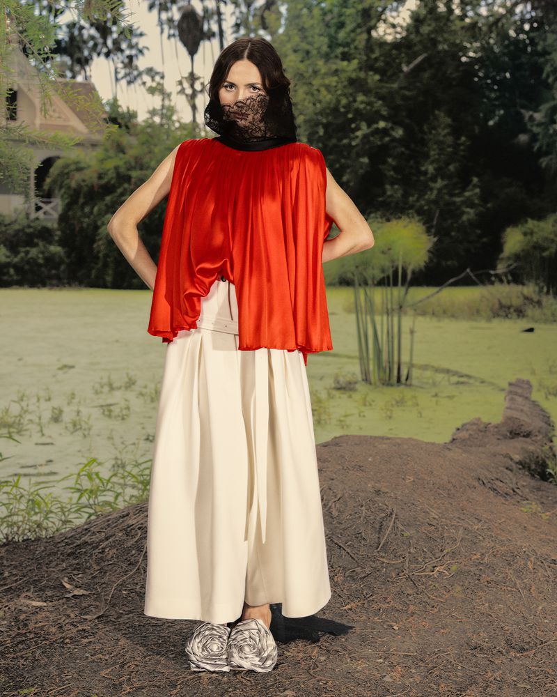 Maude Apatow wearing a red pleated top and white skirt outdoors in a garden setting for Who What Wear editorial feature