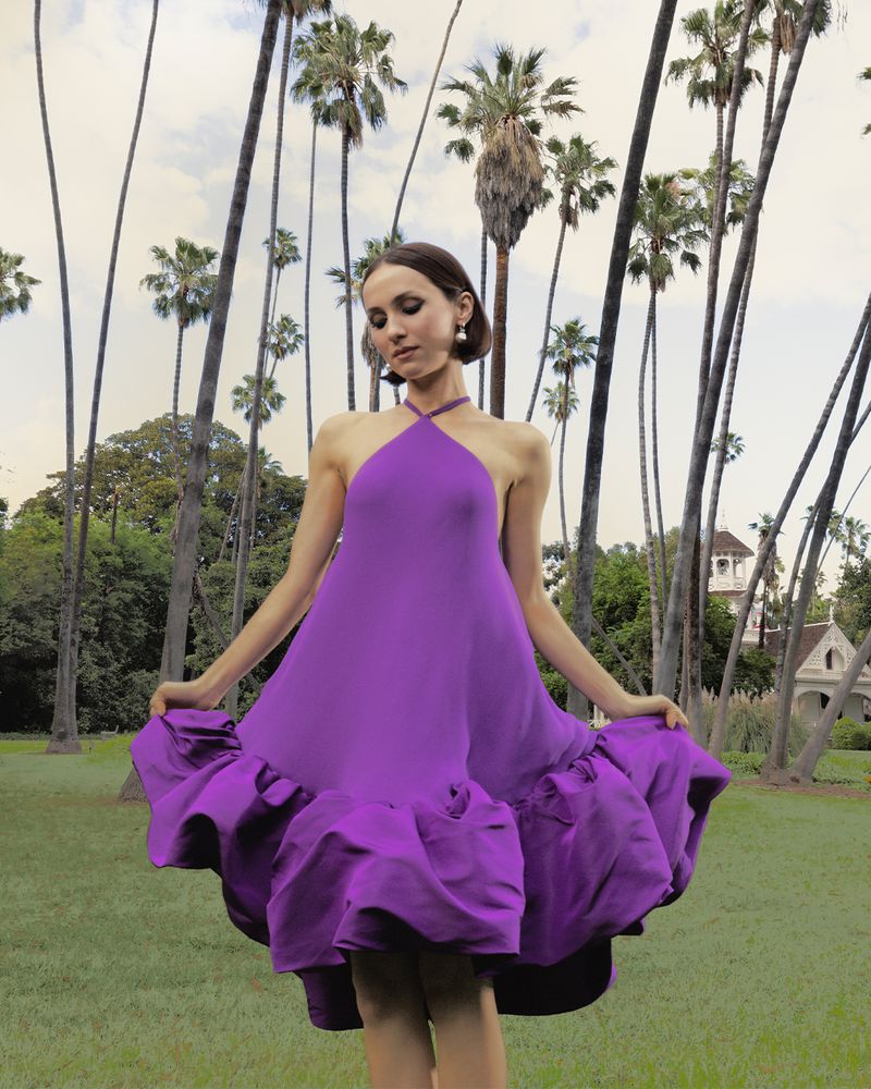 Maude Apatow wearing a vibrant purple halter minidress outdoors among tall palm trees holding the hem of the dress.