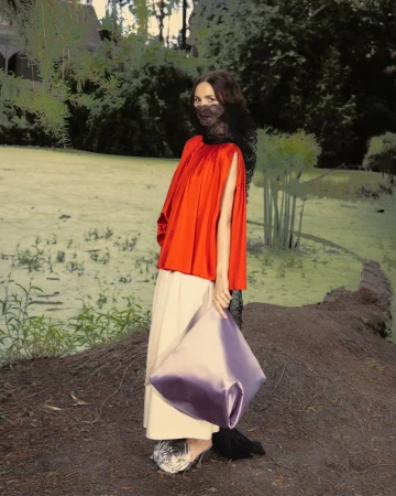 Maude Apatow in an orange top, beige skirt, and black lace veil holding a large lavender bag outdoors for Who What Wear