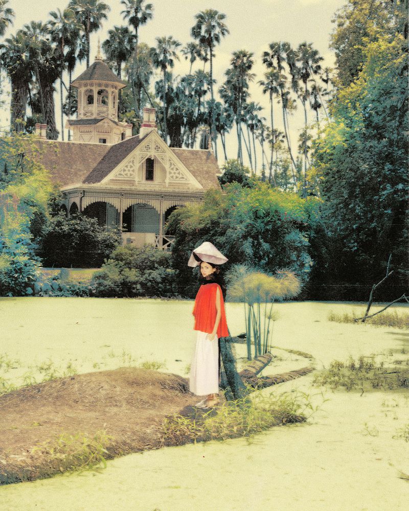 Maude Apatow standing by a pond wearing a red top and white skirt with a large hat, with a historic house and palm trees in the background.