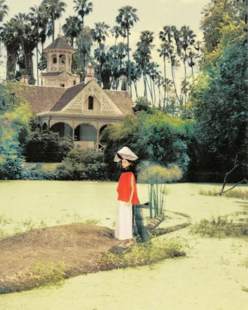 Maude Apatow standing by a pond wearing a red top and white skirt with a large hat, with a historic house and palm trees in the background.