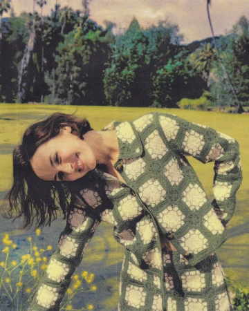 Maude Apatow posing outdoors wearing a green patterned crochet cardigan against a lush park setting.