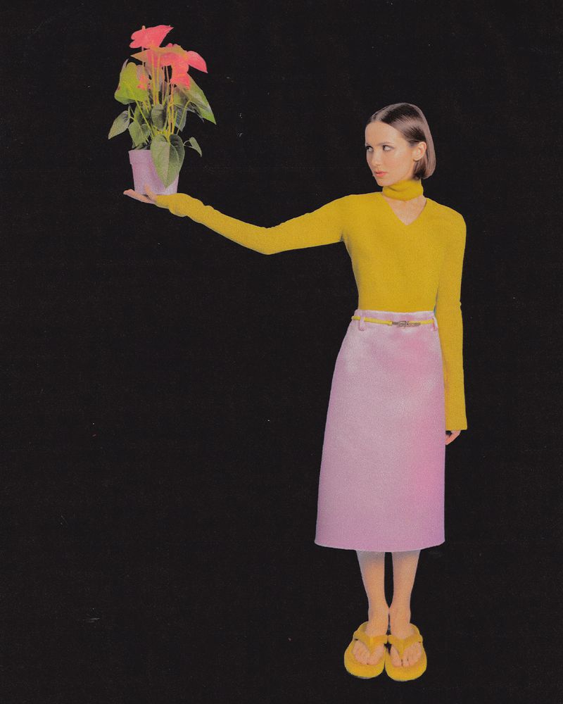 Maude Apatow holding a potted pink anthurium plant against a plain black background, wearing a yellow top and pink skirt