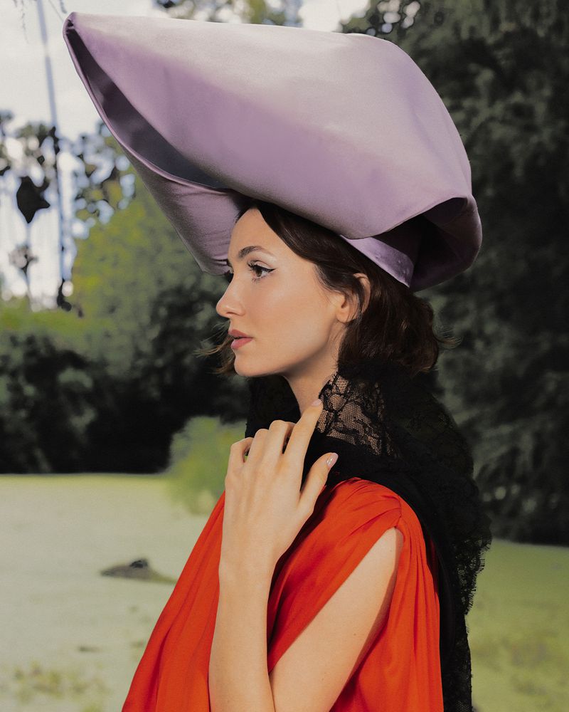 Maude Apatow in profile wearing a large purple sculptural hat, black lace scarf, and red dress against an outdoor park background.