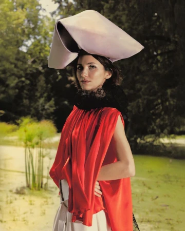 Maude Apatow wearing a red top and structured sculptural hat in an outdoor park setting for Who What Wear