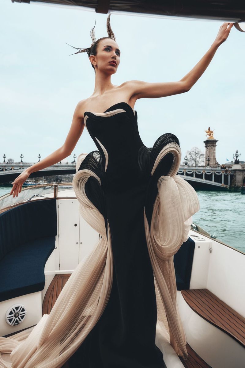 Woman in an elegant black-and-white couture gown stands on a boat with a Parisian bridge and the Seine River in the background.