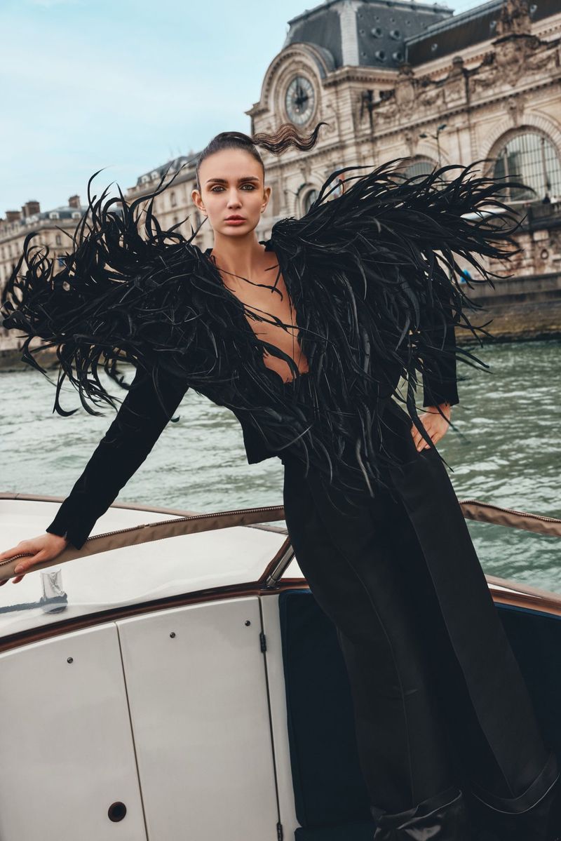 Woman poses on boat in black feather jacket and pants with city buildings in the background, creating an elegant fashion photo.