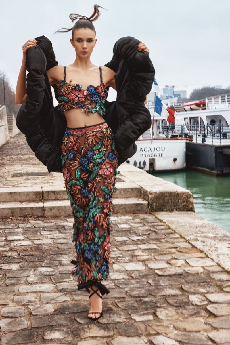 Fashion editorial shot of a woman wearing a floral two-piece outfit and black jacket, posing on a stone walkway near a river in Paris.